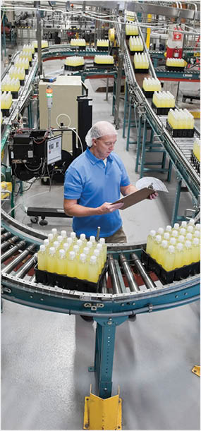 Caucasian male wearing a head net and checking inventory next to a conveyor belt of lemon flavored water in a bottling plant.